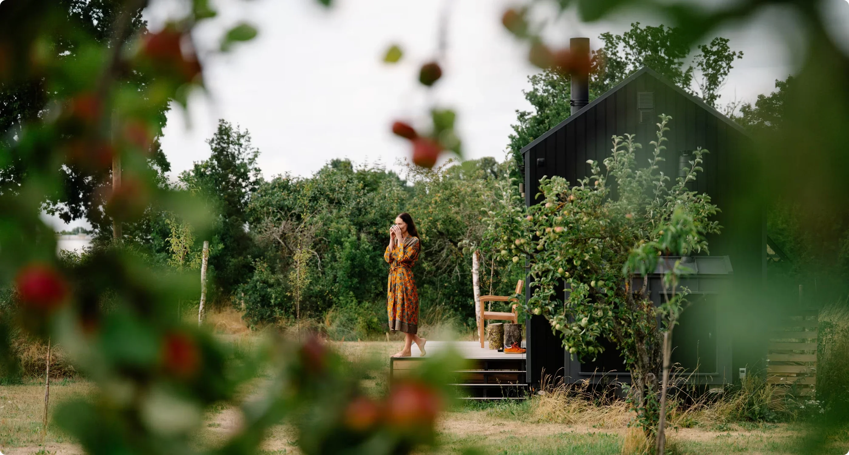 A woman on the deck of a dark wooden cabin in an orchard