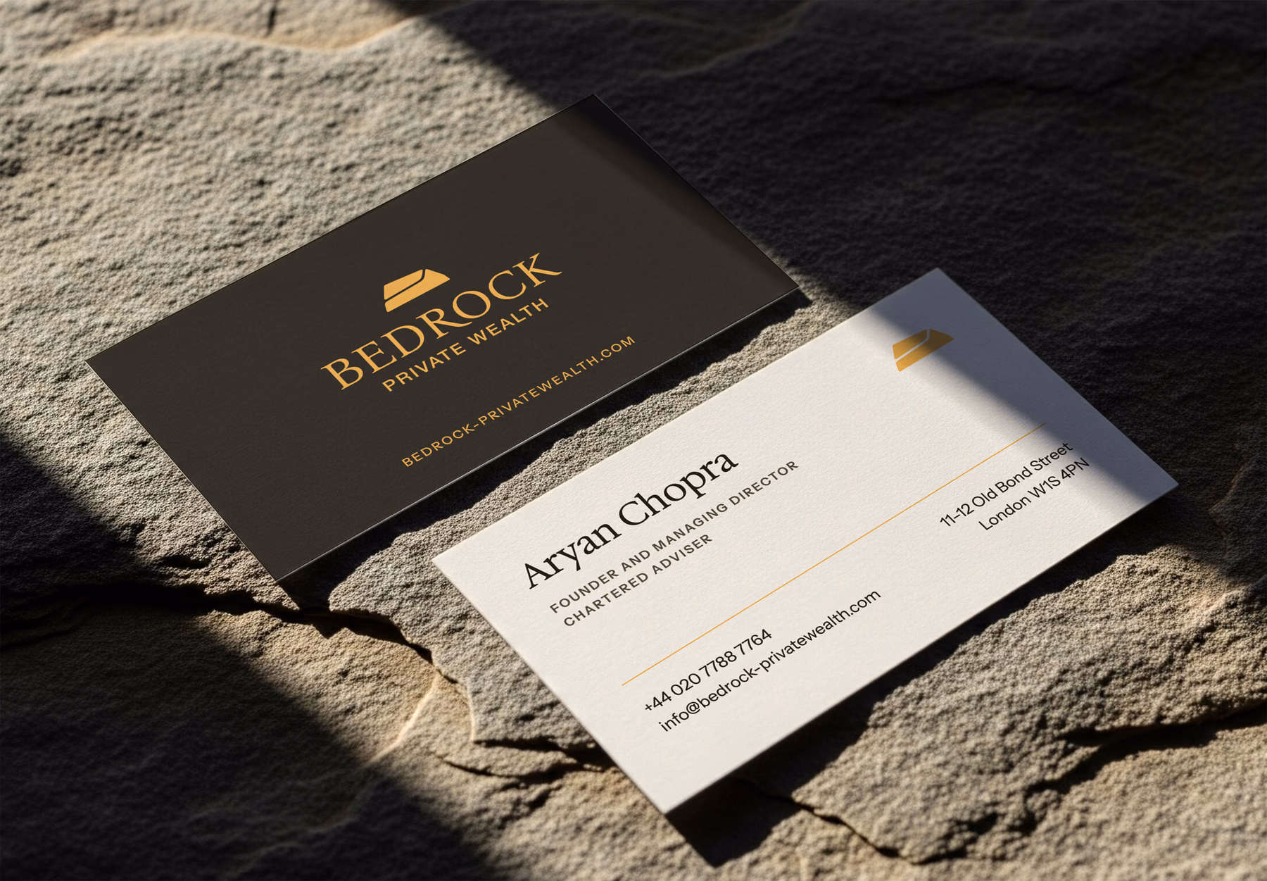 Two Bedrock Private Wealth business cards on a sunlit rock: the gold rock mark and wordmark on deep brown, and Aryan Chopra's card on warm paper with his title and the firm's contact details.